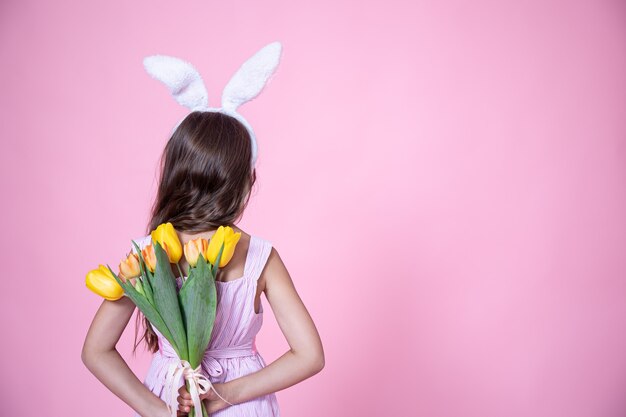 A little girl with easter bunny ears holds a bouquet of tulips in her hands behind her back on a pink studio