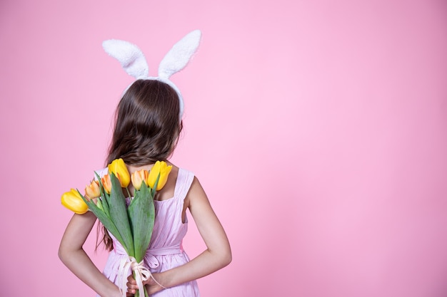 A little girl with easter bunny ears holds a bouquet of tulips in her hands behind her back on a pink studio
