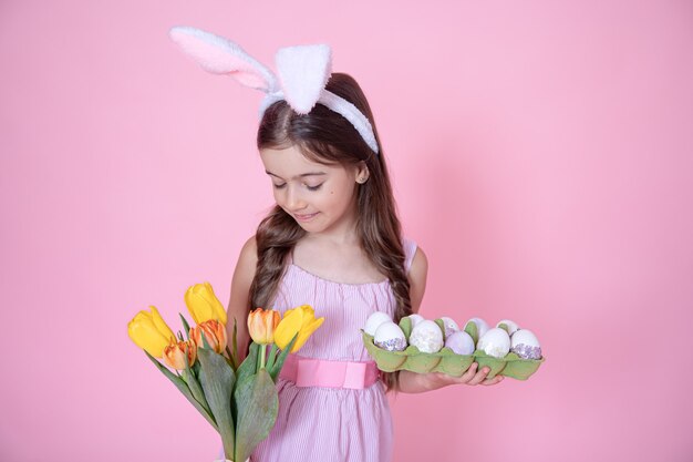 Little girl with easter bunny ears holds a bouquet of tulips and a tray of eggs in her hands on a pink wall.