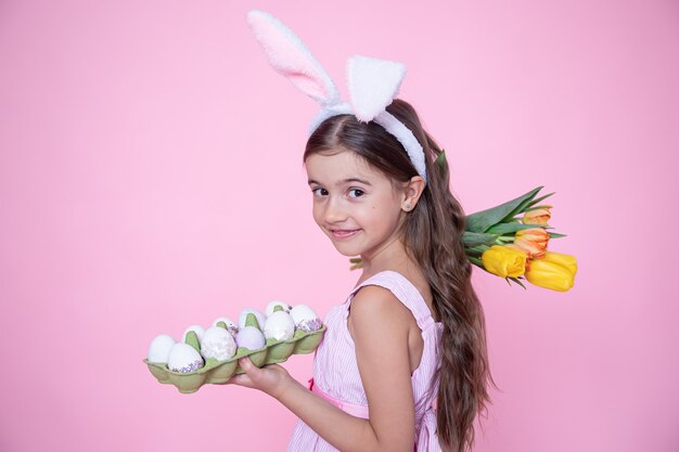 Little girl with easter bunny ears holds a bouquet of tulips and a tray of eggs in her hands on a pink wall.