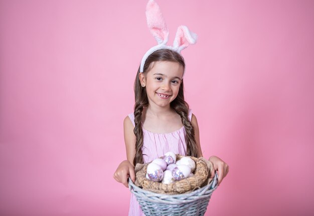 Маленькая девочка с ушками пасхального кролика позирует, держа корзину с праздничными пасхальными яйцами в розовой студии
