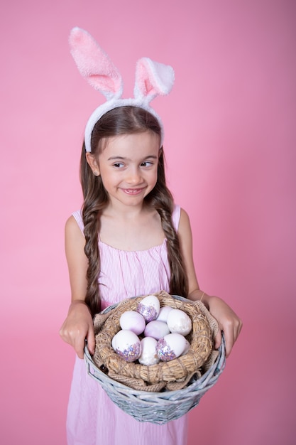 Little girl with Easter bunny ears posing holding a basket with festive Easter eggs on a pink wall close up.