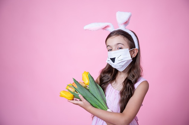 Маленькая девочка с ушками пасхального кролика и в медицинской маске держит в руках букет тюльпанов на розовой стене.
