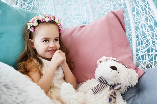 Little girl with flower wreath on her dark blonde hair holds toy white bear