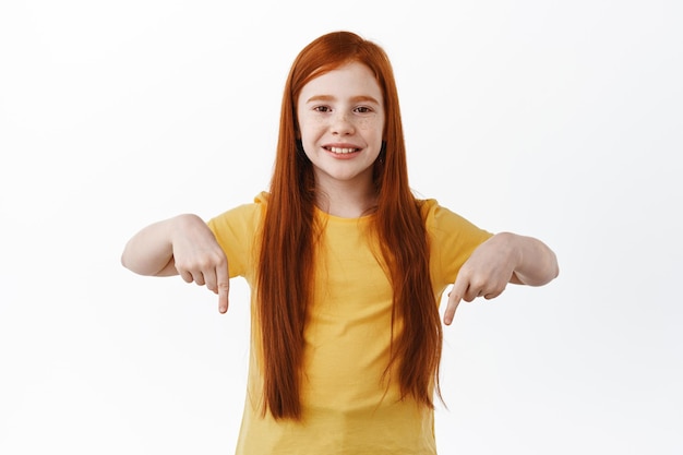 Little girl with freckles and red long hair points down. redhead kid shows logo, pointing fingers aside at copy space, smiling happy at camera, white background