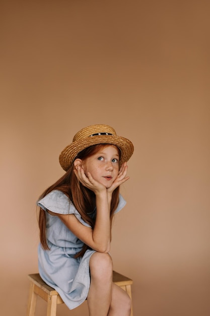 Little girl with ginger long hair and cute freckles in straw stylish hat and blue dress sitting on chair on beige background