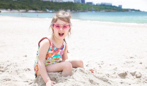 A little girl with glasses is playing in the sand on the beach by the sea.