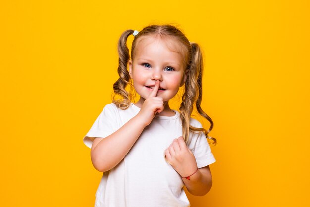 Little girl with her finger over mouth saying Shh isolated over yellow wall