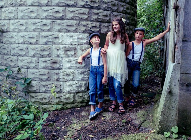 Little girl with two brothers posing near a building surrounded by greenery under sunlight