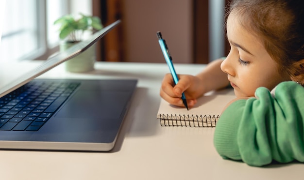 A little girl writes on a notebook while sitting in front of a laptop