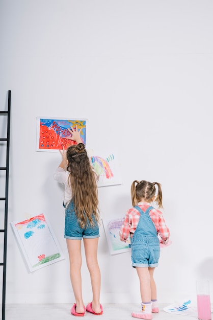 Little girls hanging drawings on white wall