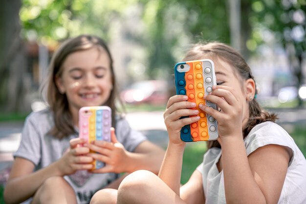 야외에서 어린 소녀들이 여드름이 있는 케이스에 휴대전화를 들고 있는데, 이것은 최신 유행의 스트레스 방지 장난감입니다.