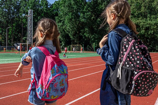 スタジアムでバックパックを背負って、男の子がサッカーをするのを見ている小さな女の子の学校の子供たち。