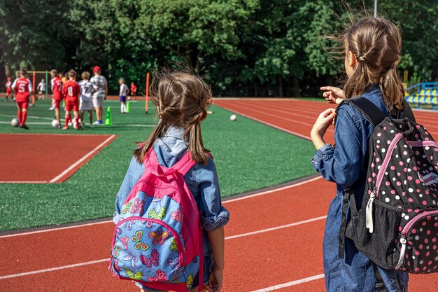 スタジアムでバックパックを背負って、男の子がサッカーをするのを見ている小さな女の子の学校の子供たち。