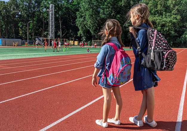 スタジアムでバックパックを背負って、男の子がサッカーをするのを見ている小さな女の子の学校の子供たち。