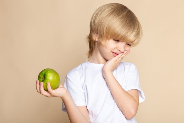 little kid blonde cute in white t-shirt holding green apple on pink wall