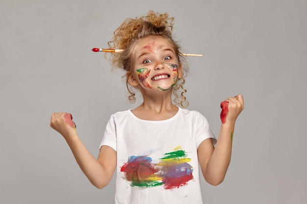 Free photo little kid having a brush in her lovely haircut, wearing in a white smeared t-shirt. she is posing with a painted hands and cheeks, making faces and looking at the camera, on a gray background.