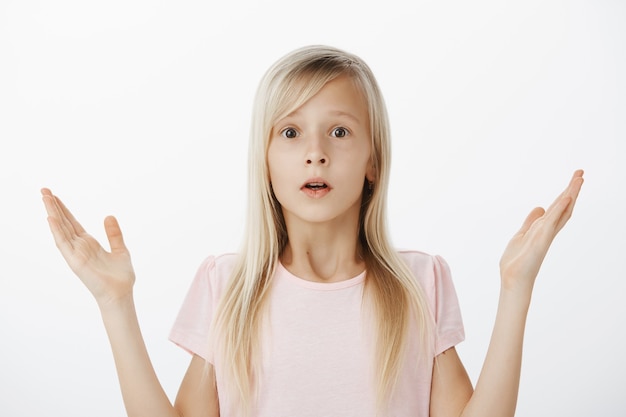 Little kid do not know anything, clueless and unaware how to act in difficult situation. Indoor shot of troubled worried cute girl with blonde hair, shrugging and raising palms, having no clue