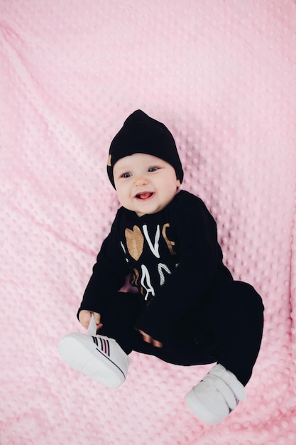 Little kid with cute big eyes lying on pink blanket on ground Sweet and lovely child with big eyes looking at camera Stylish infant in total black clothes and hat with gold heart leaning on hands