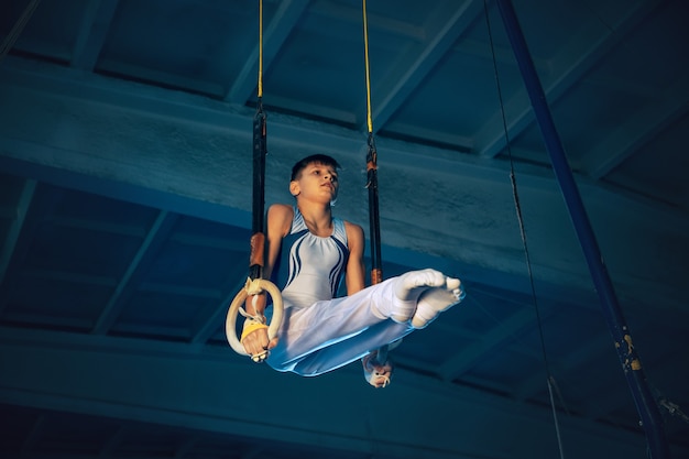 Foto gratuita piccolo ginnasta maschio allenamento in palestra, flessibile e attivo. ragazzo in forma caucasica, atleta in abiti sportivi bianchi che praticano esercizi per l'equilibrio sugli anelli. movimento, azione, movimento, concetto dinamico.