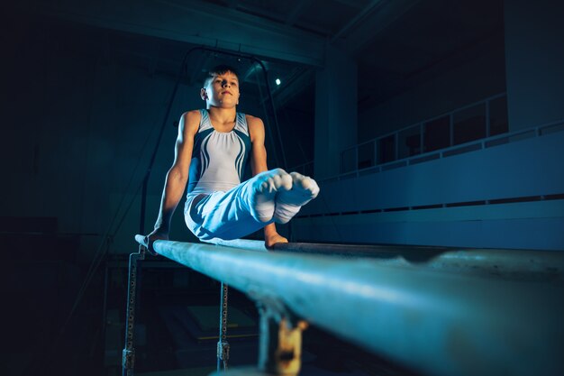 Маленький гимнастка тренируется в тренажерном зале, гибкий и активный. Кавказский маленький мальчик, спортсмен в белой спортивной одежде, упражнения на силу, баланс. Движение, действие, движение, динамическая концепция.