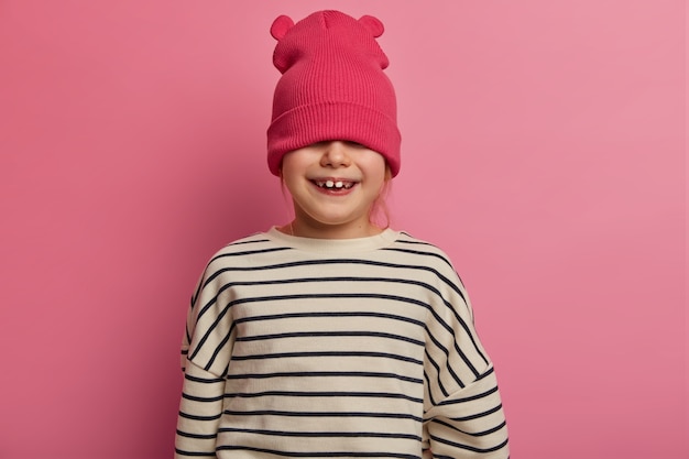 Little naughty girl hides eyes with stylish hat, has fun and doesnt want go to kindergartne, has toothy smile, being in great upbeat mood, poses over pastel rosy wall. Children, fashion, style