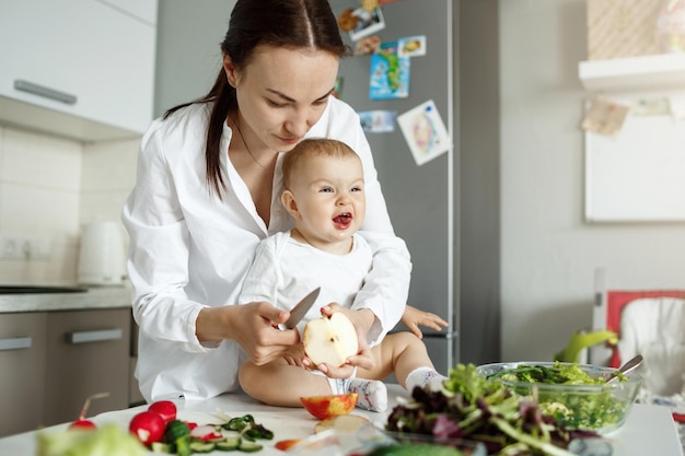 お母さんが家族の昼食のためにサラダを調理している間、おかしな表情で母親の前のテーブルに座っている小さな新生児。家族の概念。