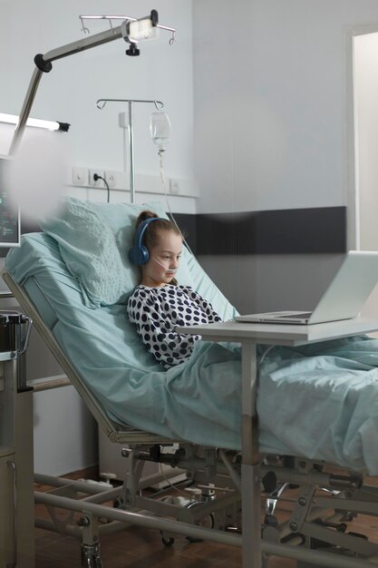 Little patient girl under treatment resting on bed in hospital pediatrics ward room while watching funny cartoons on modern laptop. Sick little kid enjoying internet video content while hospitalized.