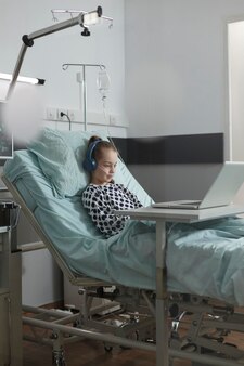 現代​の​ラップ​トップ​で​面白い​漫画​を​見​ながら​、​病院​の​小児科​病棟​の​ベッド​で​休む​治療中​の​小さな​患者​の​女の子​。​入院中​に​インターネット ビデオ コンテンツ​を​楽しんでいる​病気​の​小さな​子供​。