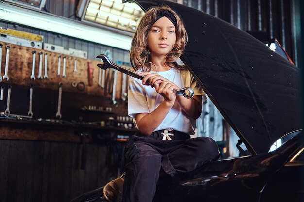 Little pensive girl is sitting on the open car's hood with big wrench in hands.