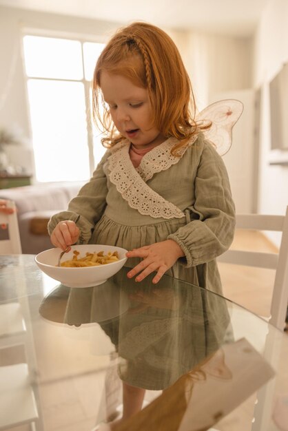 小さな赤毛の白人の子供の女の子は、テーブルに座って食事をしながらおいしい料理を楽しむ離乳食のコンセプト