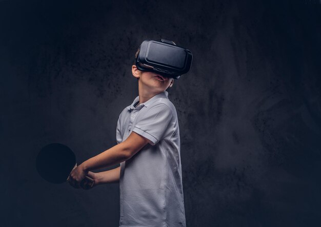 Little redhead boy dressed in a white t-shirt playing ping-pong with a virtual reality glasses. Isolated on a dark textured background.
