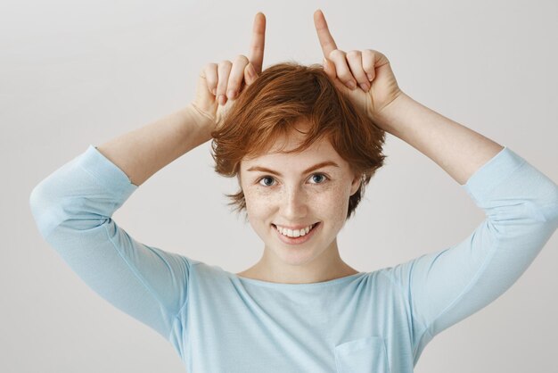 Little redhead devil makes guy fall for her from first sight Portrait of attractive european woman with ginger hair making horns on top of head with index fingers and smiling with interest