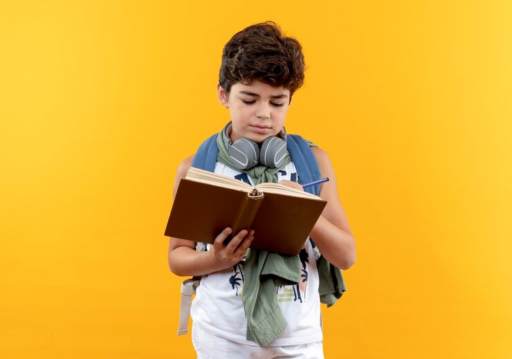 मुफ़्त फोटो little school boy wearing back bag and headphones writing something on book isolated on yellow background