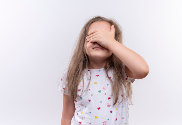 Little school girl wearing white t-shirt covered eyes with hand on isolated white wall