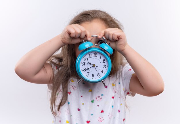  little school girl wearing white t-shirt covered face with alarm clock on isolated white wall