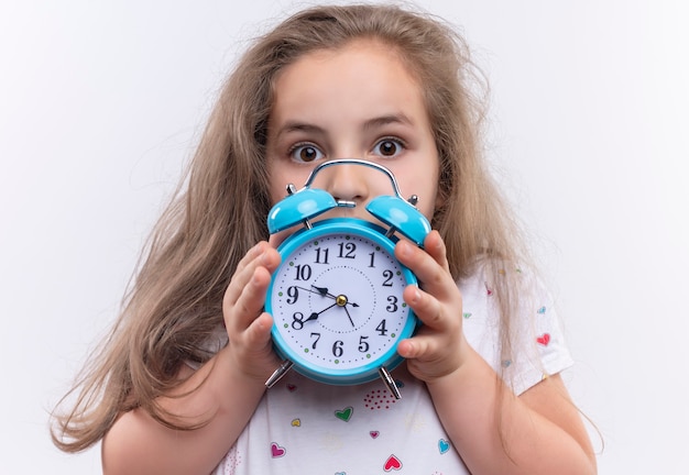  little school girl wearing white t-shirt holding alarm clock on isolated white wall