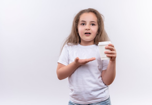 隔離の白い壁にコーヒーのカップを保持している白いTシャツを着ている小さな女子高生