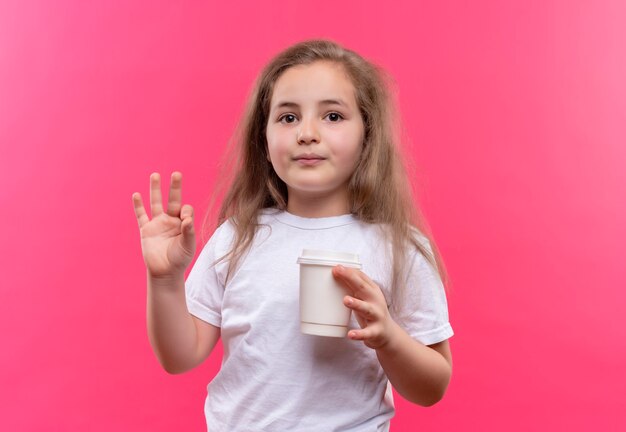 little school girl wearing white t-shirt holding cup of coffee showing okey gesture on isolated pink wall