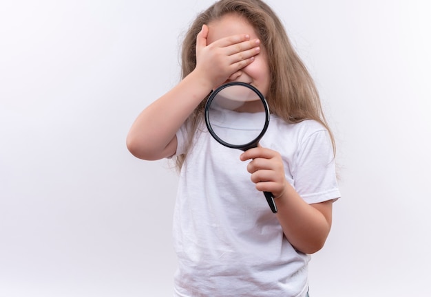 Little school girl wearing white t-shirt holding magnifier covered eyes with hand on isolated white wall