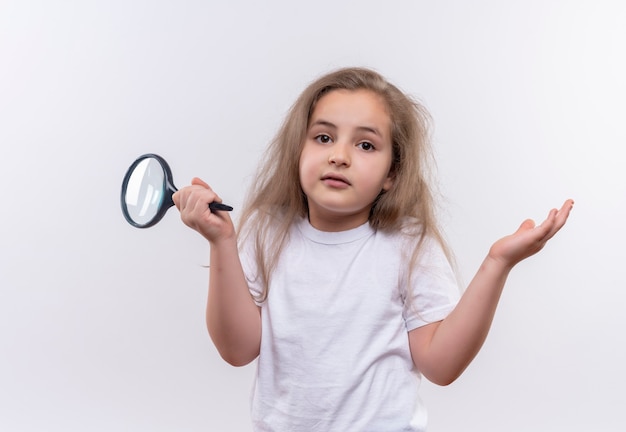 little school girl wearing white t-shirt holding magnifier showing what gesture on isolated white wall