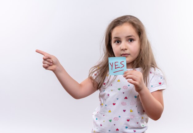  little school girl wearing white t-shirt holding paper mark points to side on isolated white wall