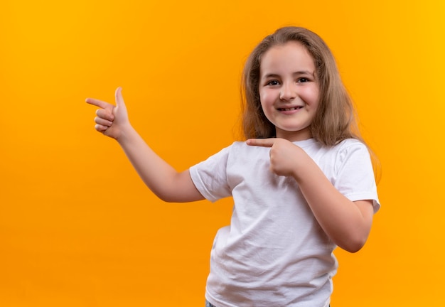 little school girl wearing white t-shirt points to side on isolated orange wall