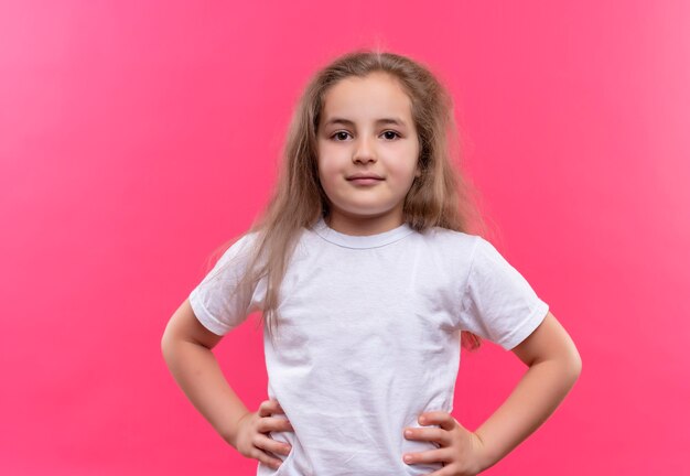 Маленькая школьница в белой футболке положила руки на бедра на изолированной розовой стене