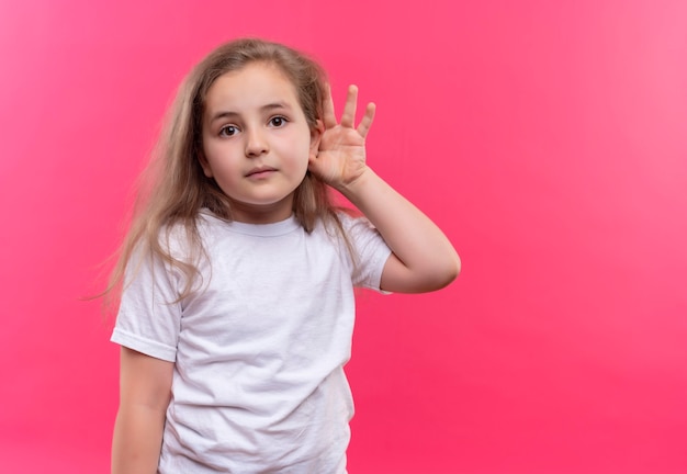 little school girl wearing white t-shirt showing listen gesture on isolated pink wall