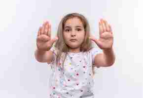 Free photo little school girl wearing white t-shirt showing stop gesture on isolated white wall