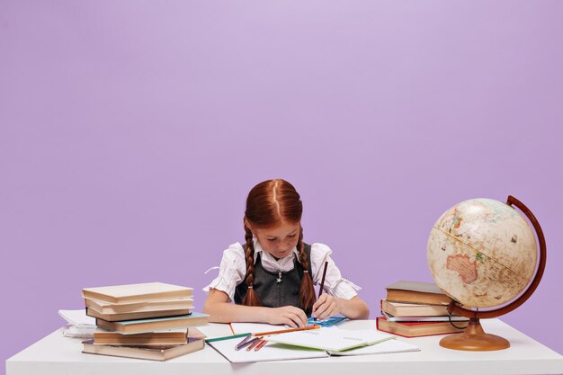 Little school girl with two red braids in white blouse and sundress sits at table with books and globe on lilac background