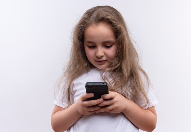 Little school woman wearing white t-shirt playing with phone on isolated white wall
