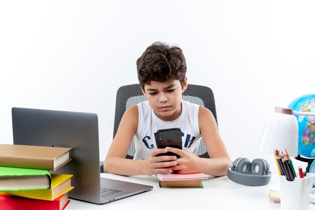 little schoolboy sitting at desk with school tools holding and looking at phone