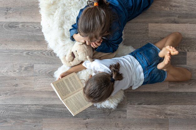 Little sisters are reading a book with a teddy bear lying on the floor in the room top view.